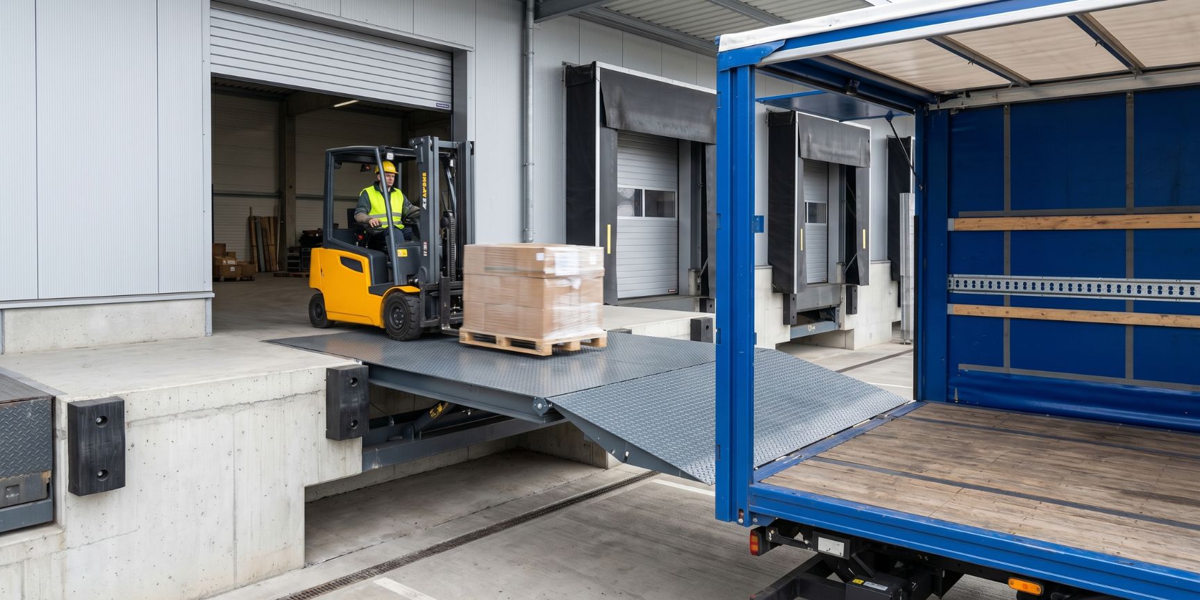Photo of a bascule wedge bridge in use. The steel platform connects a logistics ramp with a truck, and the forklift truck drives onto the loading area. Folding lip rests securely.